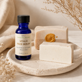 A cobalt blue glass bottle of Niki's The Natural Boutique VITALIS FACE SERUM stands on a round stone tray beside two handmade soap bars, one with a gold wax seal, in a warm neutral setting with dried botanicals.