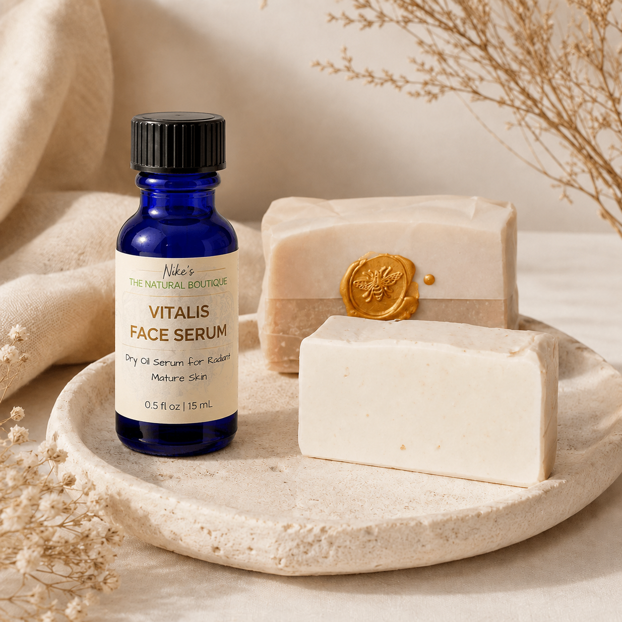 A cobalt blue glass bottle of Niki's The Natural Boutique VITALIS FACE SERUM stands on a round stone tray beside two handmade soap bars, one with a gold wax seal, in a warm neutral setting with dried botanicals.