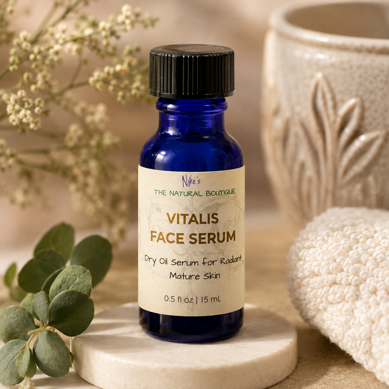 A cobalt blue glass bottle of Niko's The Natural Boutique Vitalis Face Serum with a black cap, displayed on a marble coaster with eucalyptus leaves and a textured ceramic background.